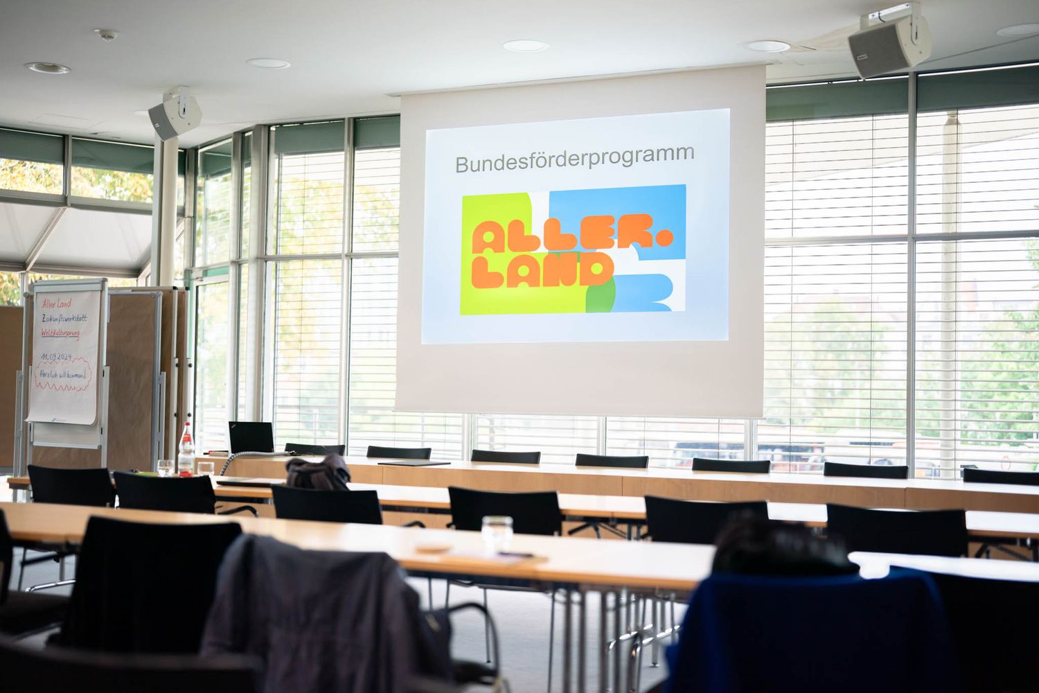 Empty classroom with rows of desks and chairs facing a whiteboard in a bright room with large windows.