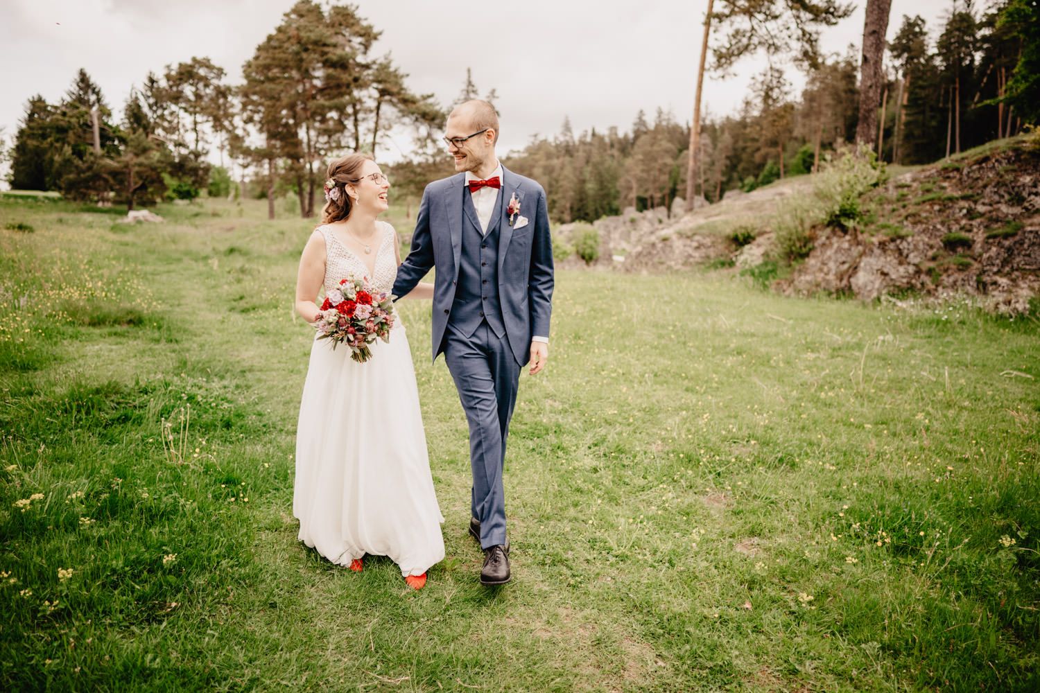 Couple walking together on grassy hillside with pine trees in background.