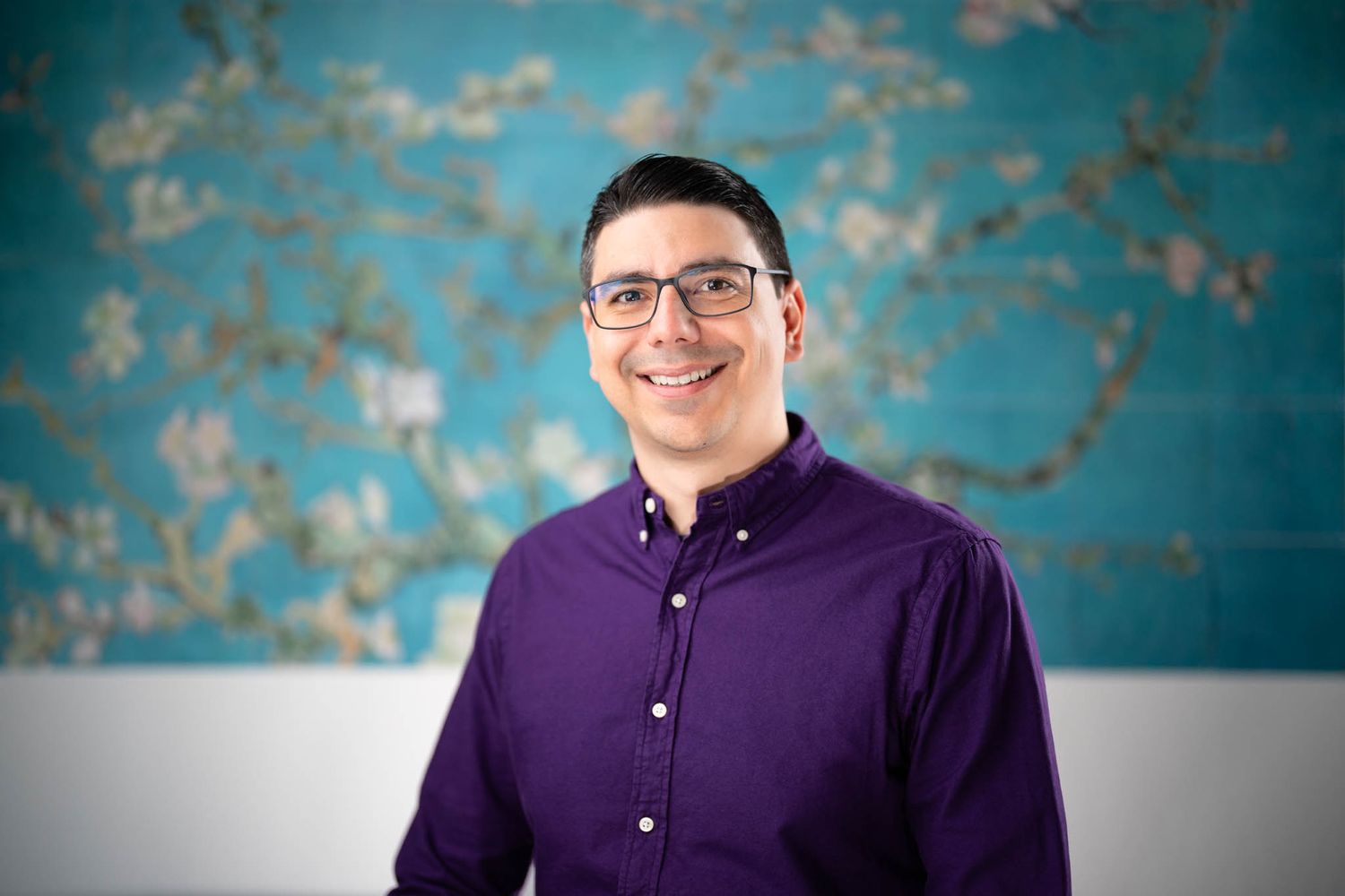 Professional portrait against a textured turquoise wall wearing a purple button-down shirt and glasses.