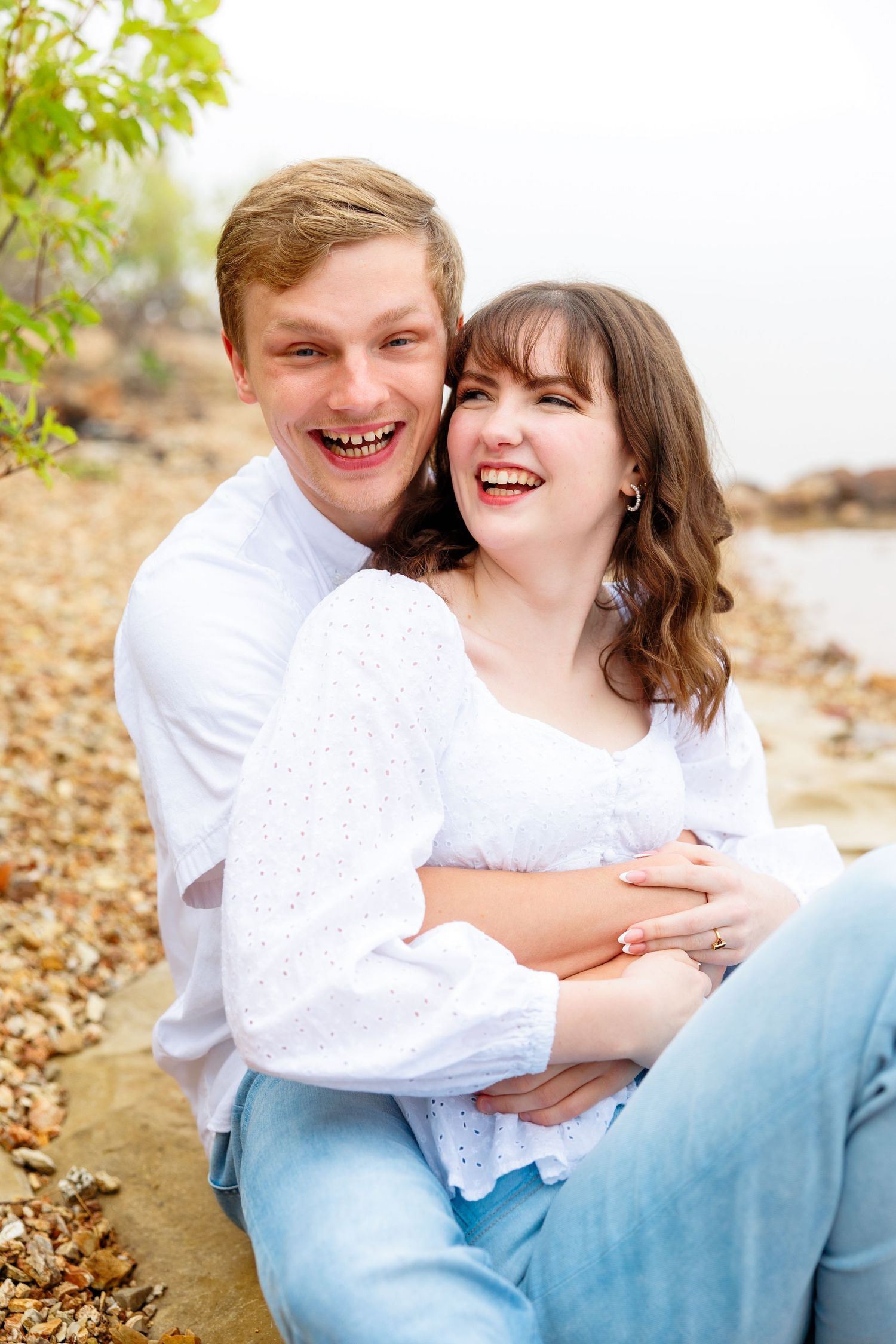 Happy couple laughing together on a rocky beach, both wearing white tops and jeans in an outdoor engagement photo in Branson, MO.