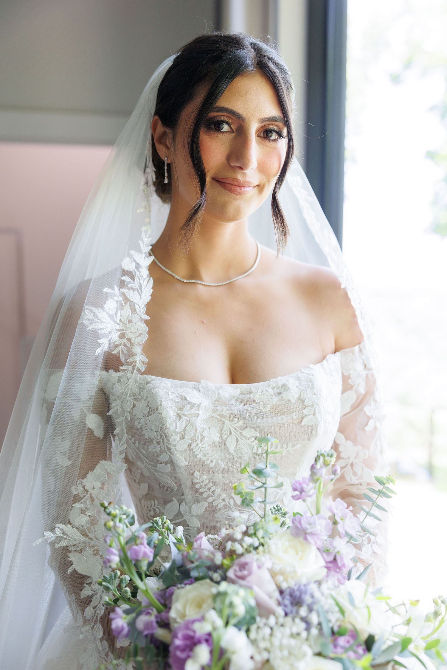 Elegant bride in lace off-shoulder gown and veil holding purple floral bouquet, smiling softly near window.