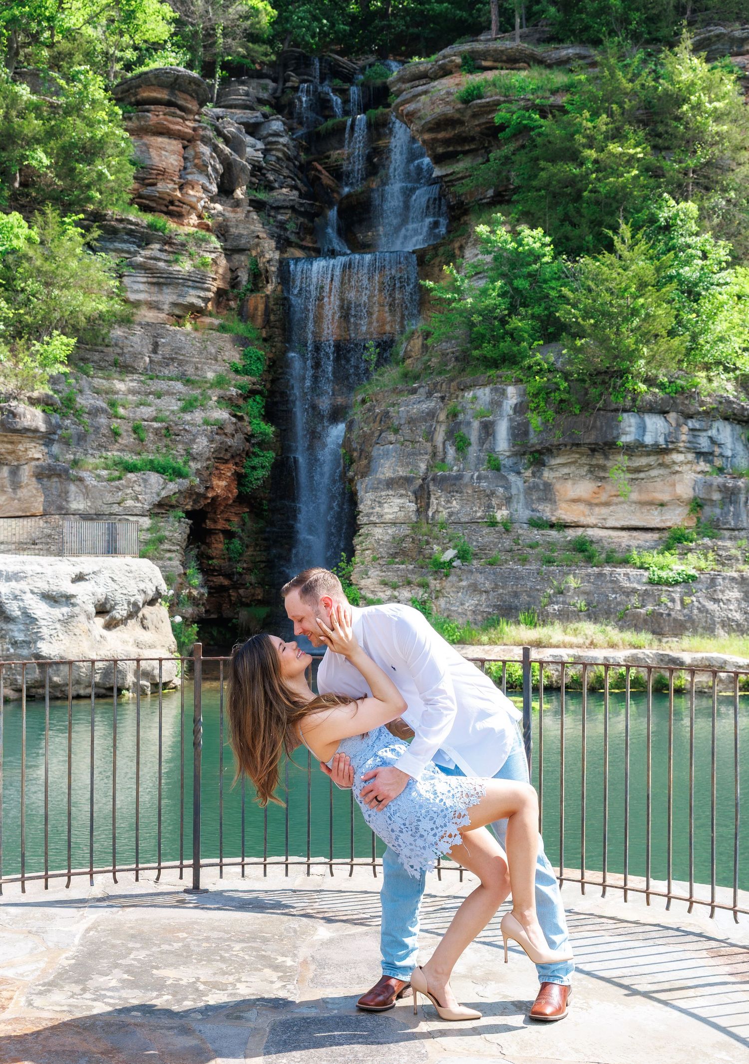 Couple sharing a romantic dip kiss in front of a stunning waterfall cascading over rocky cliffs into a green pool.