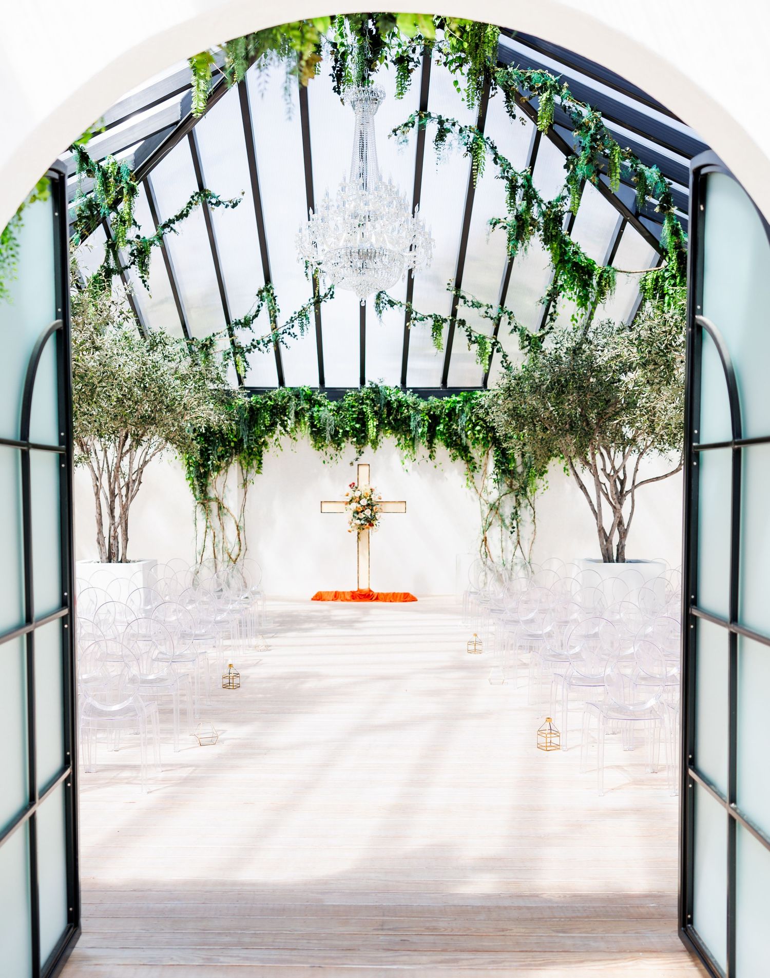 Elegant greenhouse wedding chapel with glass ceiling, lush greenery, crystal chandelier, white chairs, and a gold cross altar.