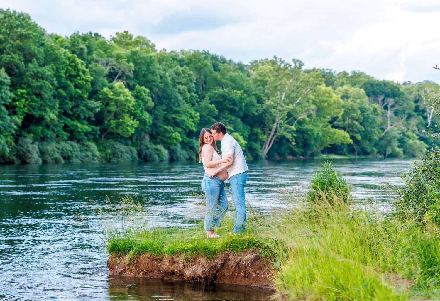 Couple sharing a romantic kiss on a lush riverbank surrounded by green trees and calm water.