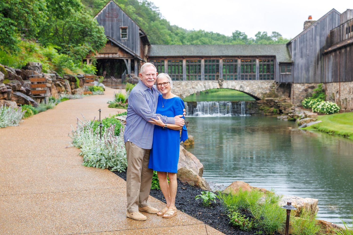 Couple posing together by a scenic pond with a stone bridge, waterfall, and covered wooden bridge in a lush garden setting.