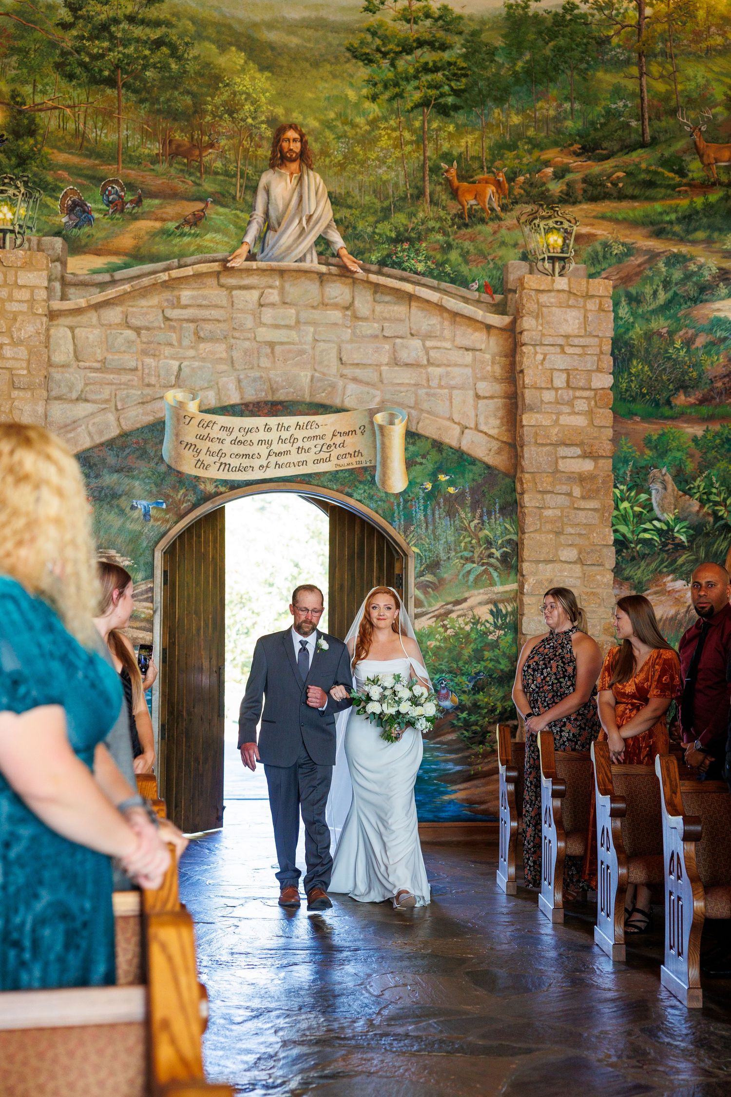A wedding ceremony processional takes place in a stone archway corridor decorated with scenic wall murals.