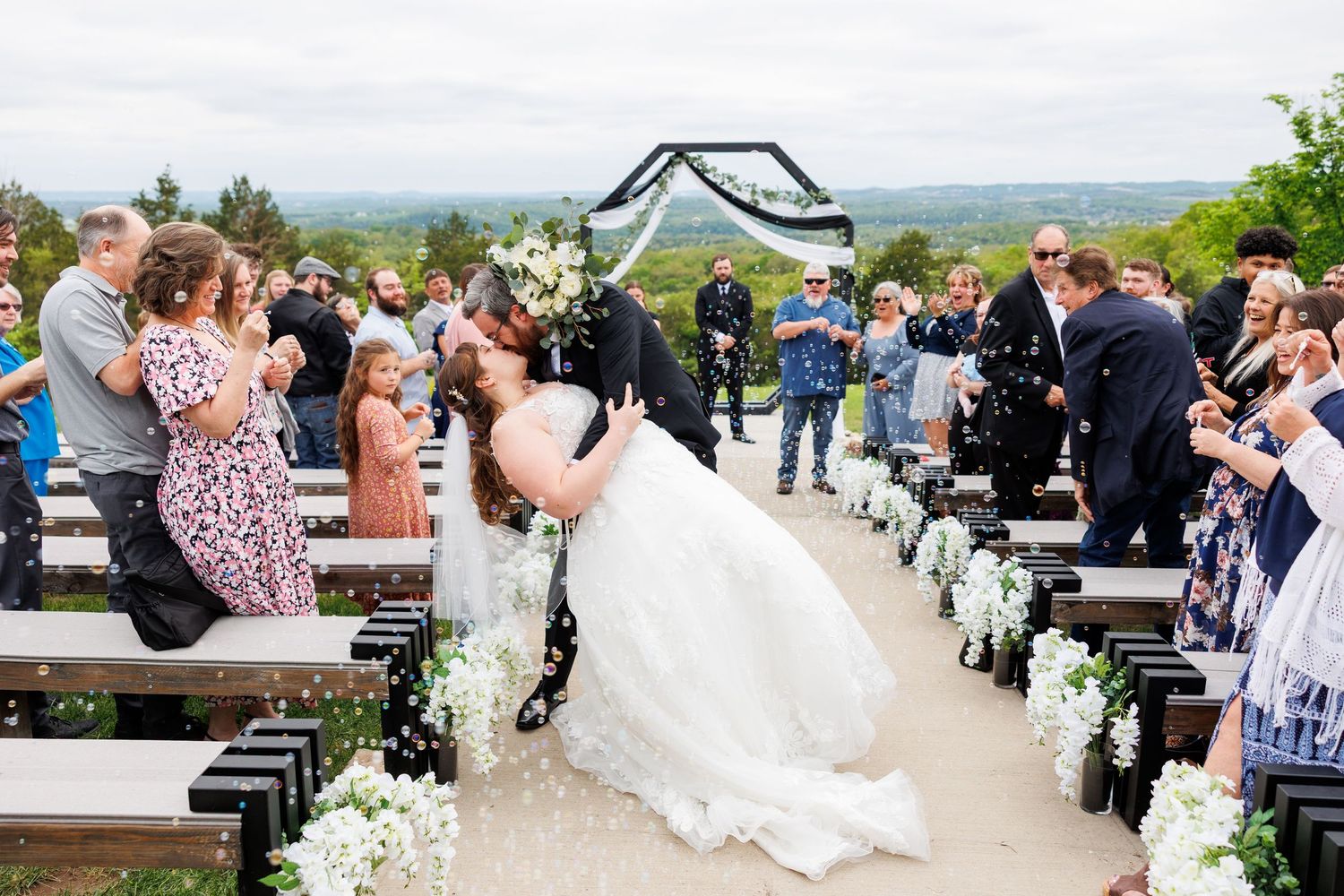 Groom dips bride for a kiss at outdoor wedding ceremony while guests cheer along flower-lined aisle at the Atrium in Branson, MO.