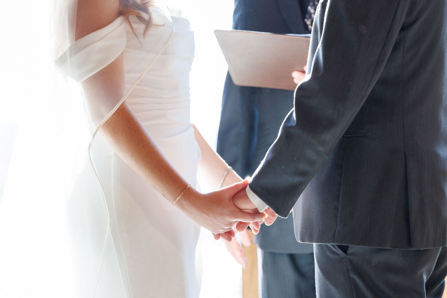 A couple holds hands during a wedding ceremony, with the bride wearing white and groom in a dark suit.