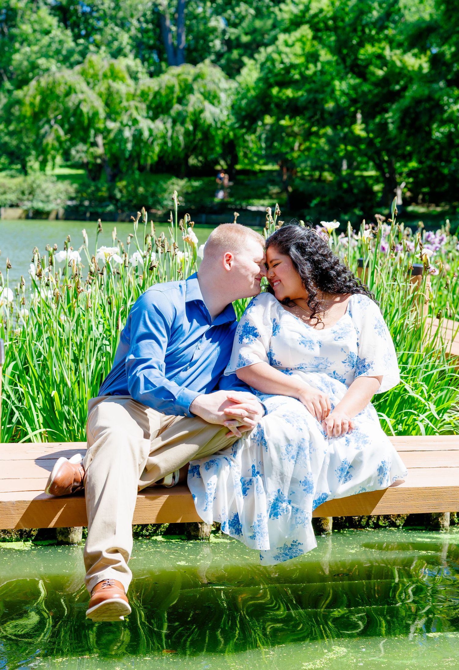 Couple sharing a romantic moment on a dock surrounded by iris flowers and a lush green pond on a sunny day.