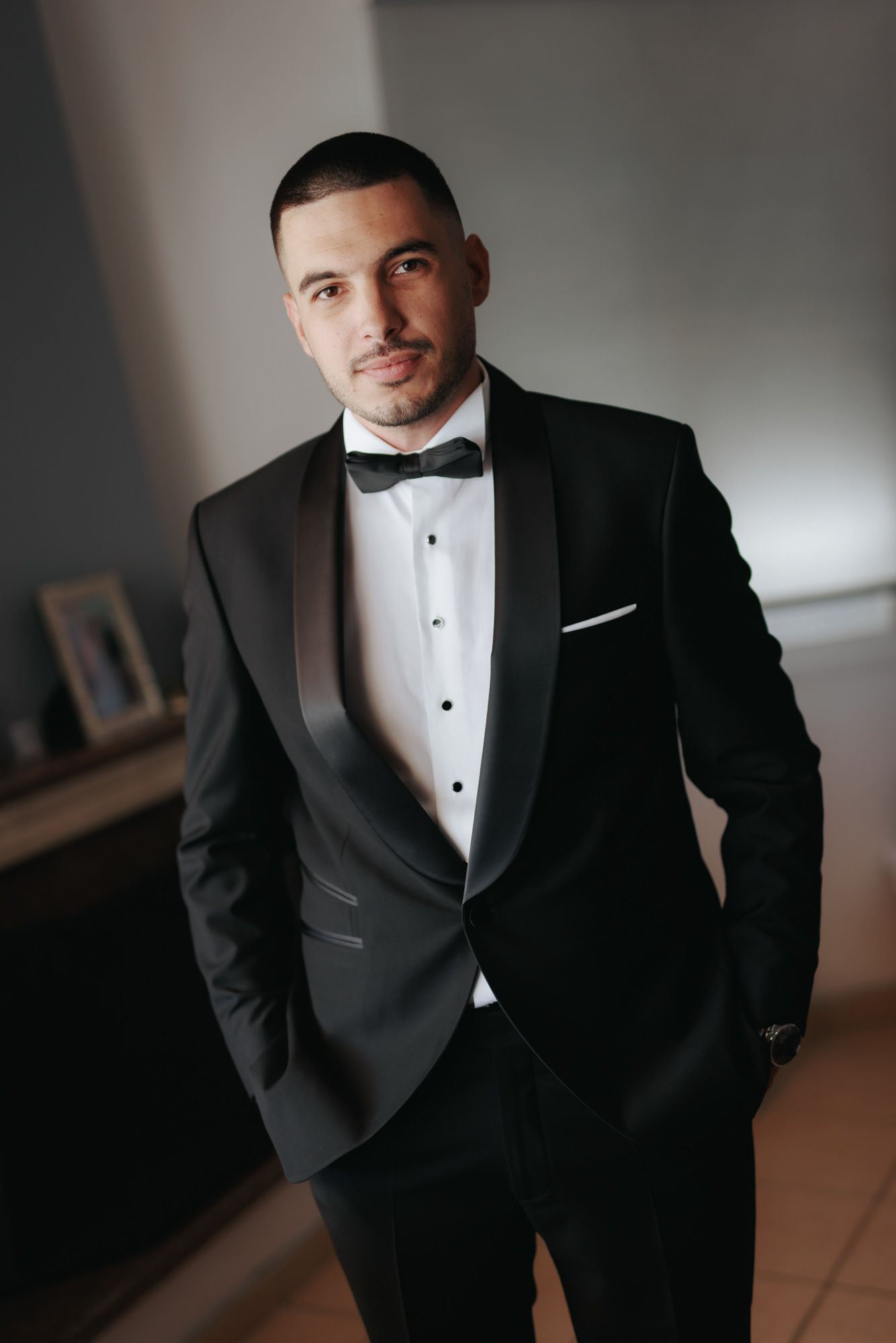 A well-dressed groom in a black tuxedo with bow tie and white pocket square poses confidently indoors.