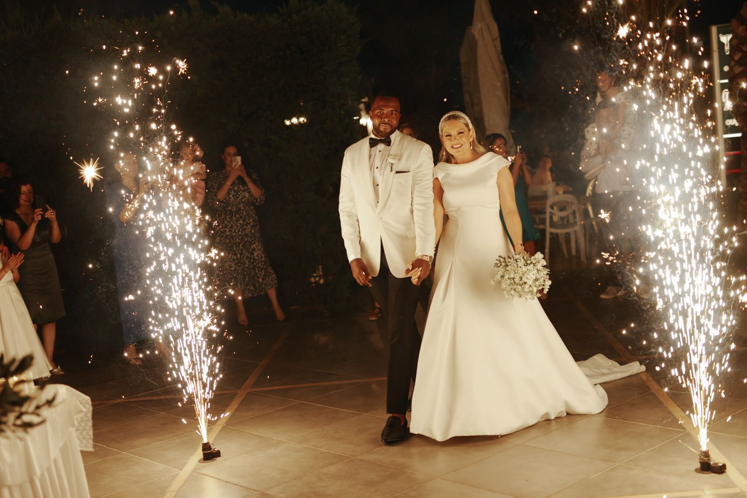 Bride and groom walk through sparkling wedding fireworks display at elegant outdoor evening reception ceremony.