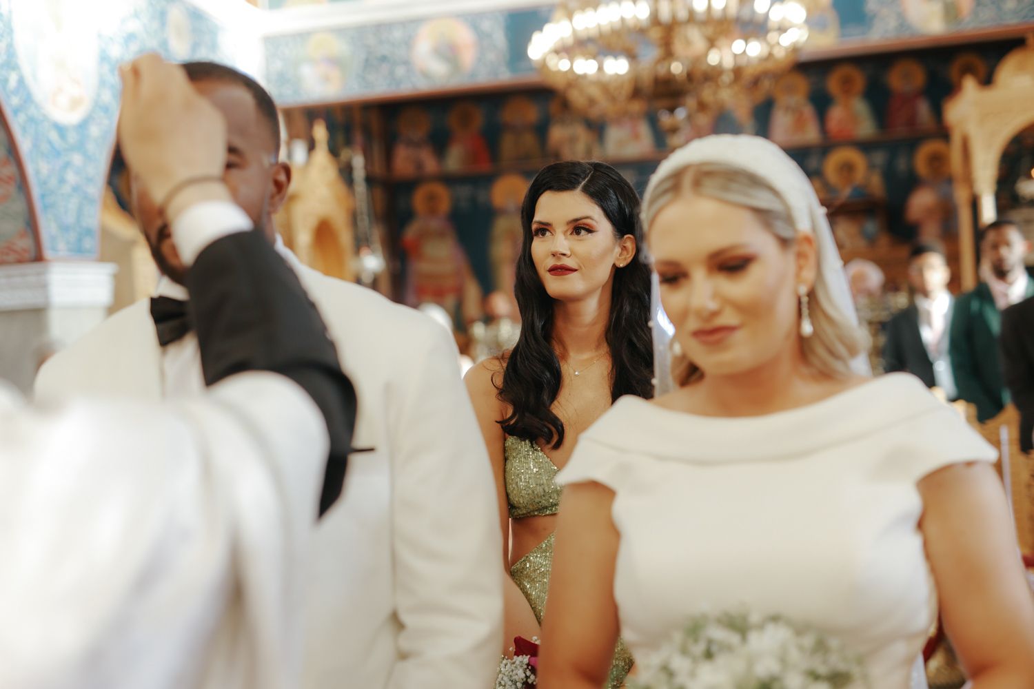 Bride in white gown during Orthodox church wedding ceremony with guests in elegant attire in background.