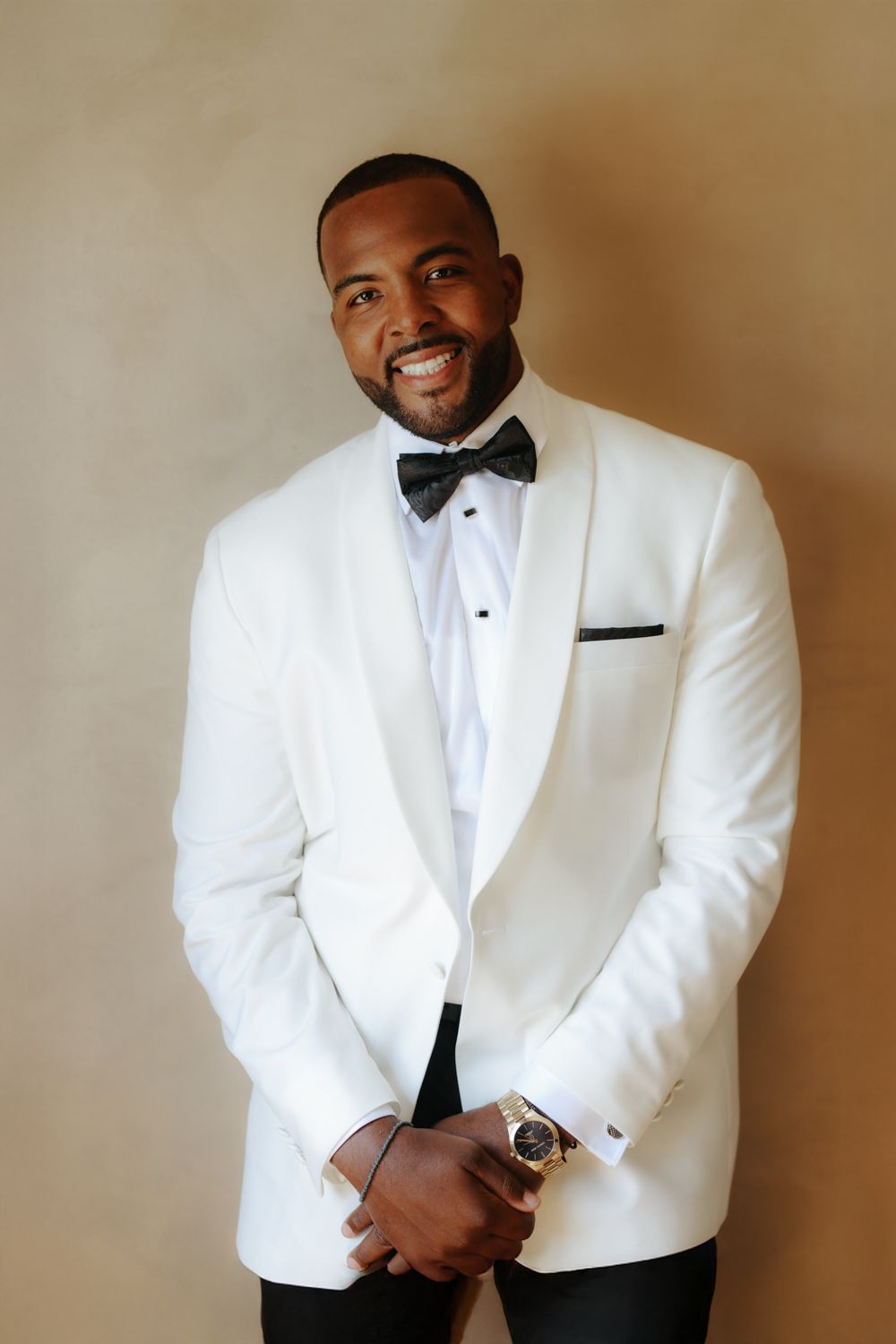 Smiling man in white tuxedo jacket and black bow tie posing confidently against a neutral background.