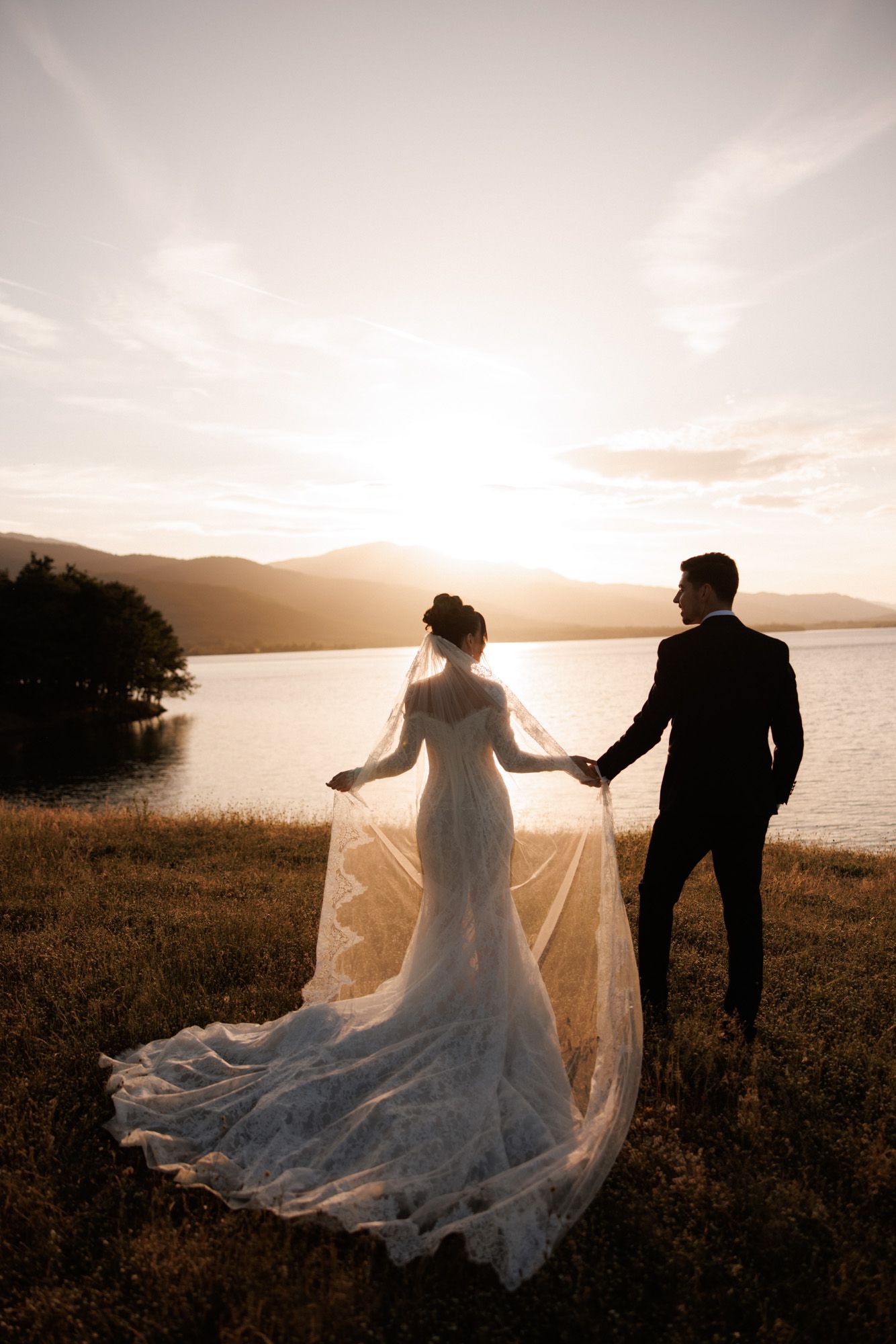 Bride and groom holding hands at sunset by a lake, with her lace wedding gown train flowing in the breeze.