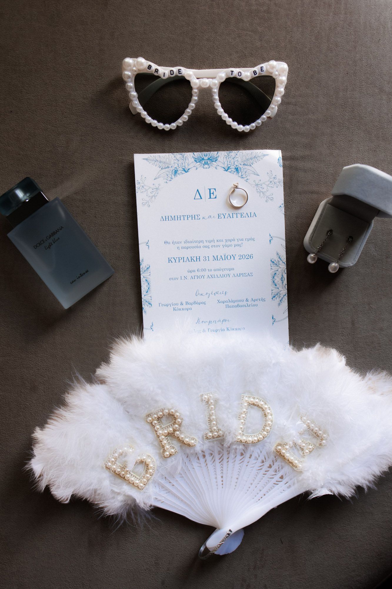 Bridal flat lay with feather fan, pearl heart sunglasses, wedding invitation, rings, and perfume bottles on gray background.