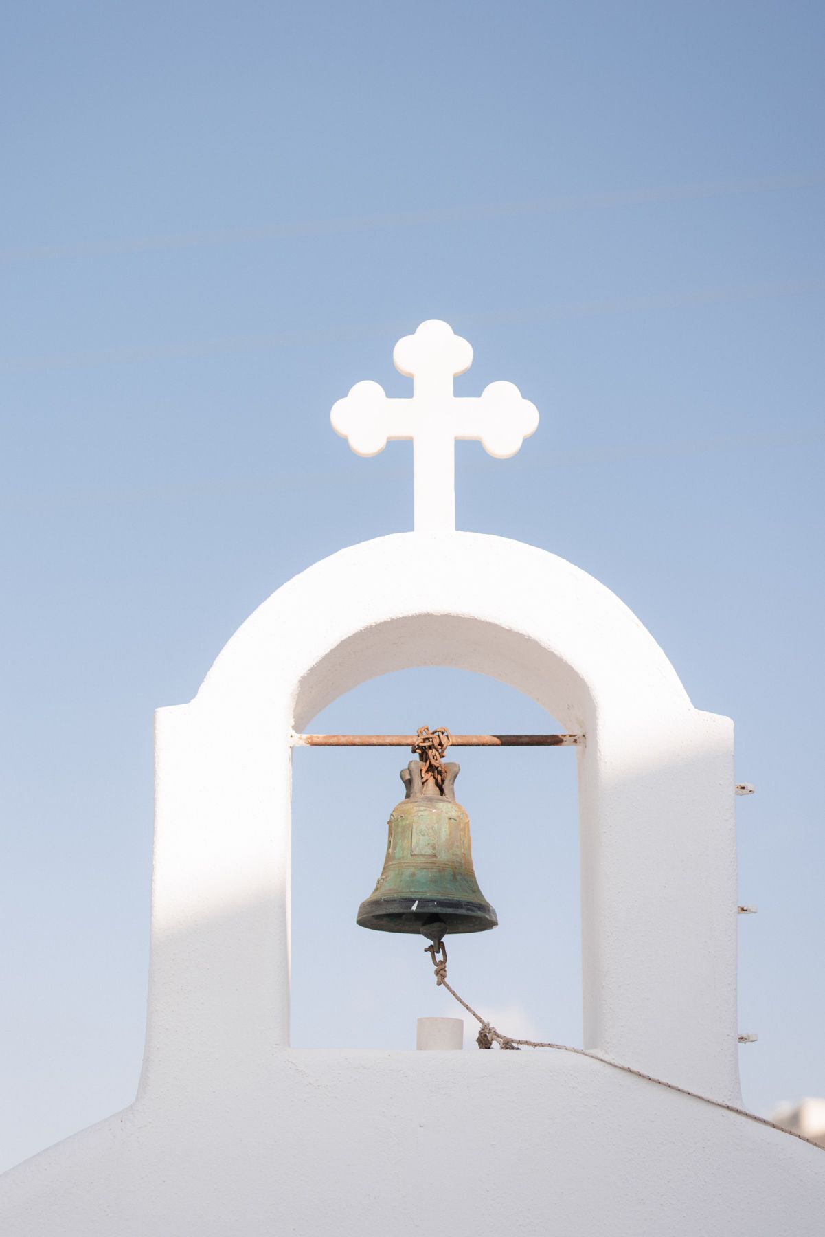 Traditional white Greek Orthodox church bell tower with bronze bell against clear blue sky.