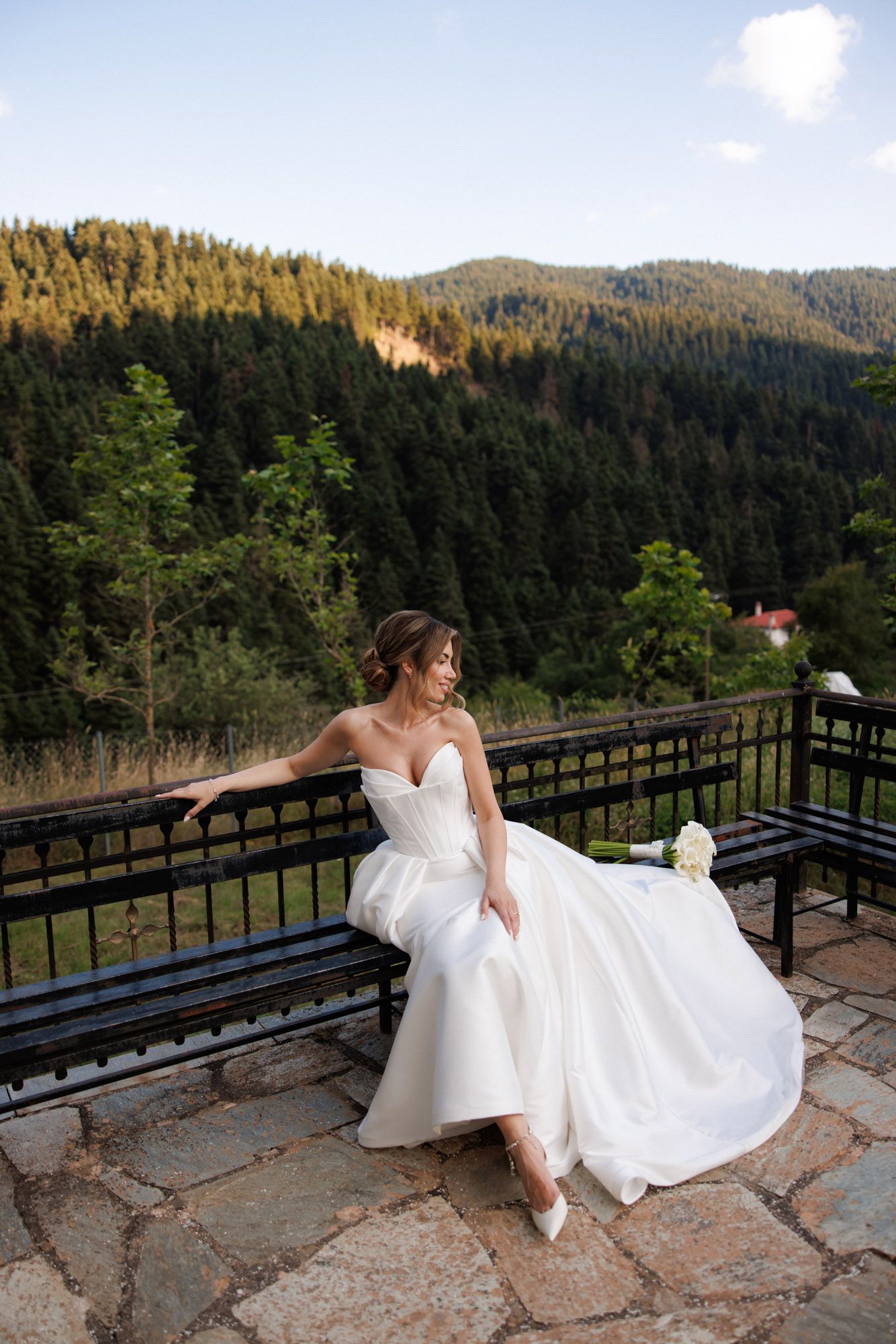Bride in strapless white gown sits on bench with stunning forested mountain landscape behind her at dusk.