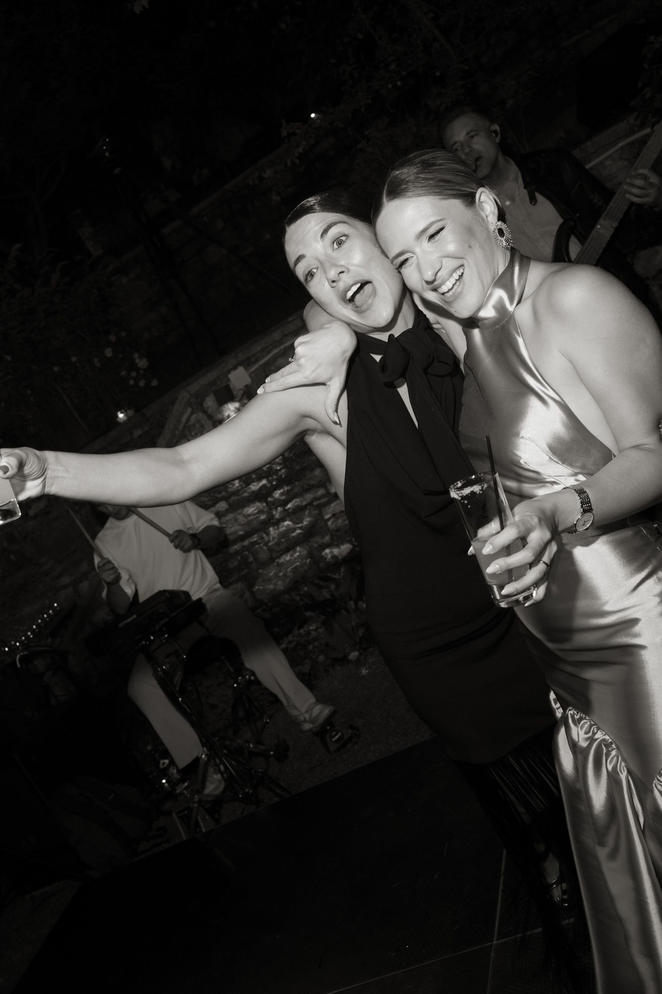Two women in formal attire laughing and dancing together at wedding reception in black and white.
