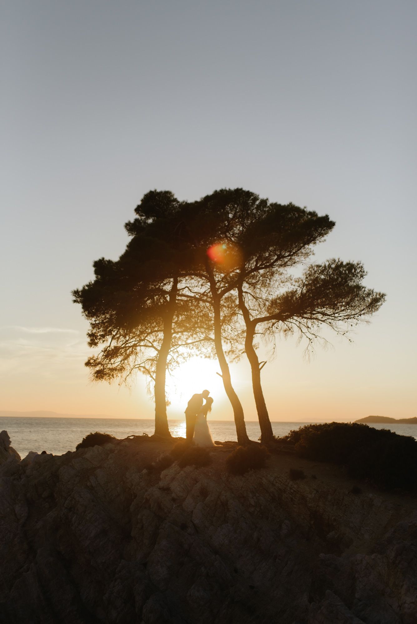 Romantic couple silhouetted under pine trees on rocky coast during stunning golden sunset by the sea.