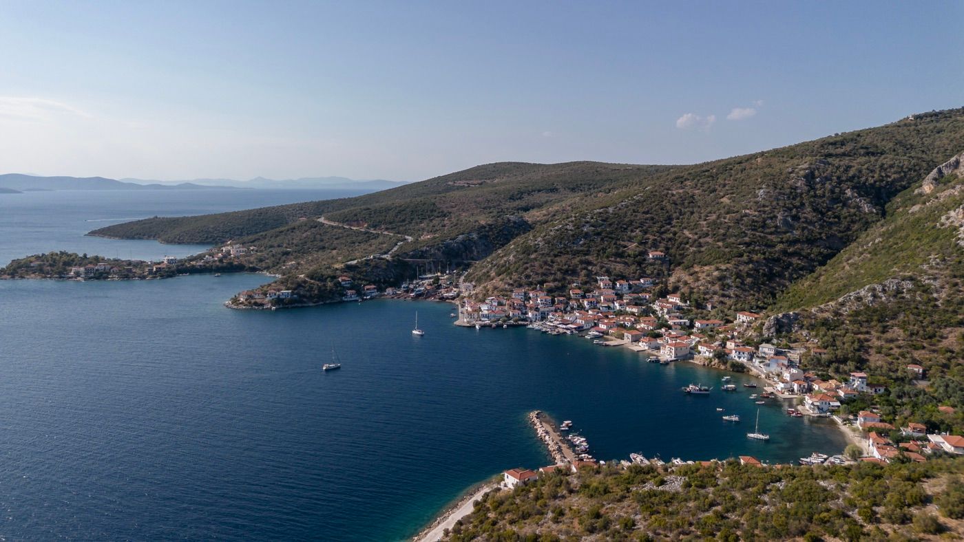 Aerial panoramic view of a scenic coastal town with marina and lush green hillside surroundings.