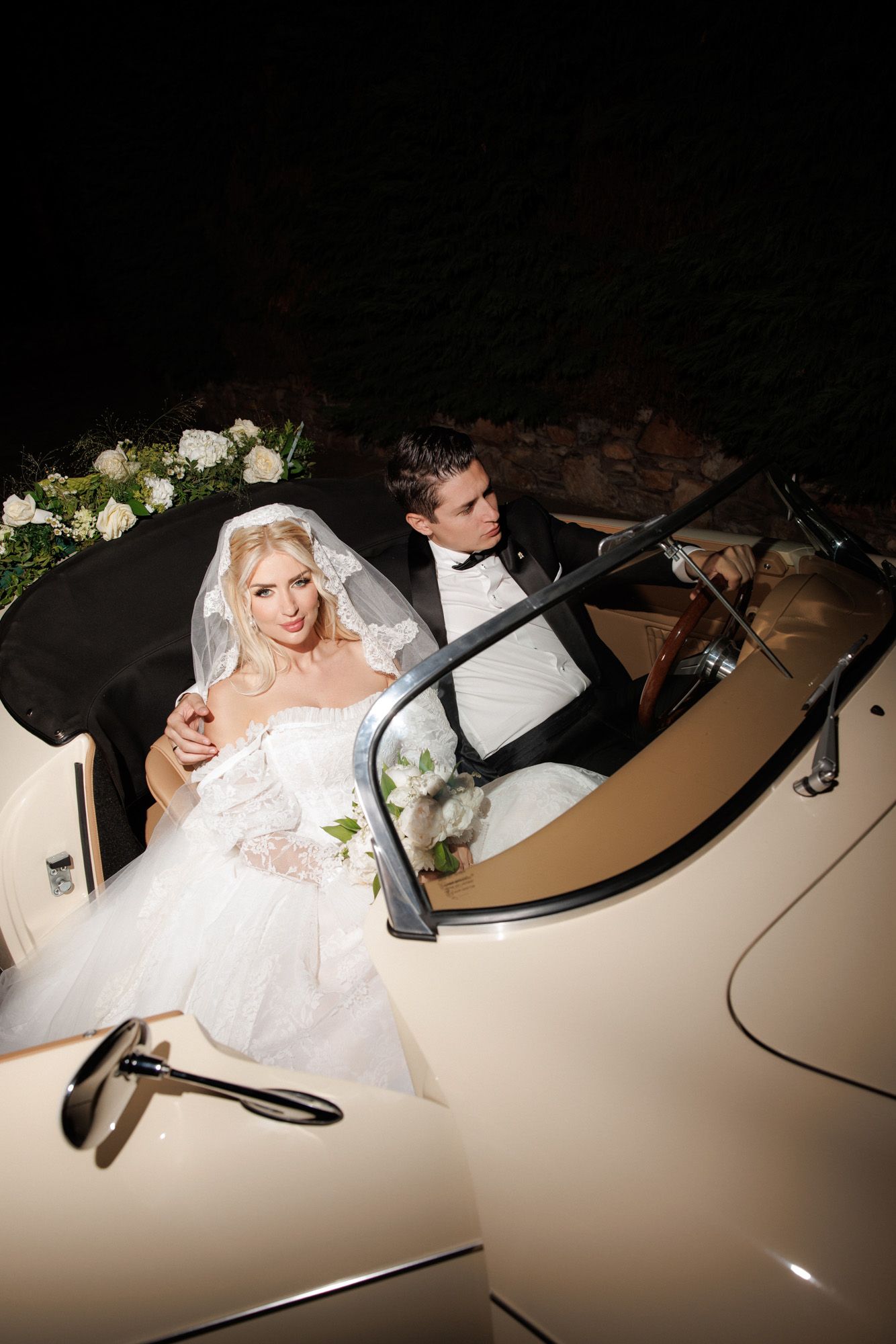 Bride and groom seated in vintage cream convertible car at night wedding.