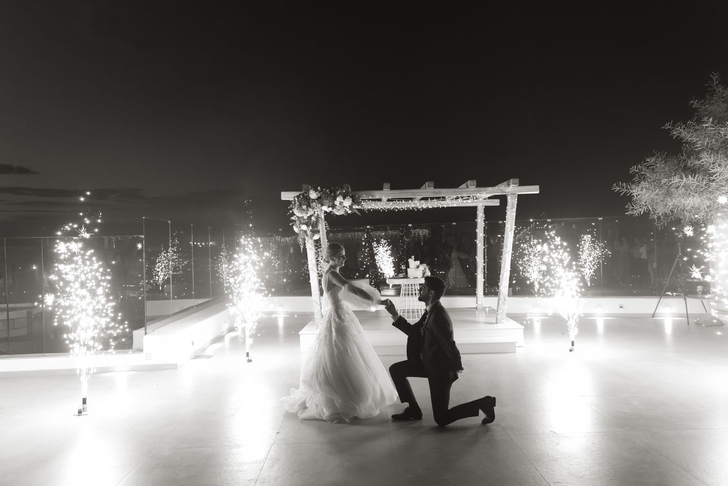 Groom kneels before bride during romantic first dance with pyrotechnic sparklers at outdoor wedding.