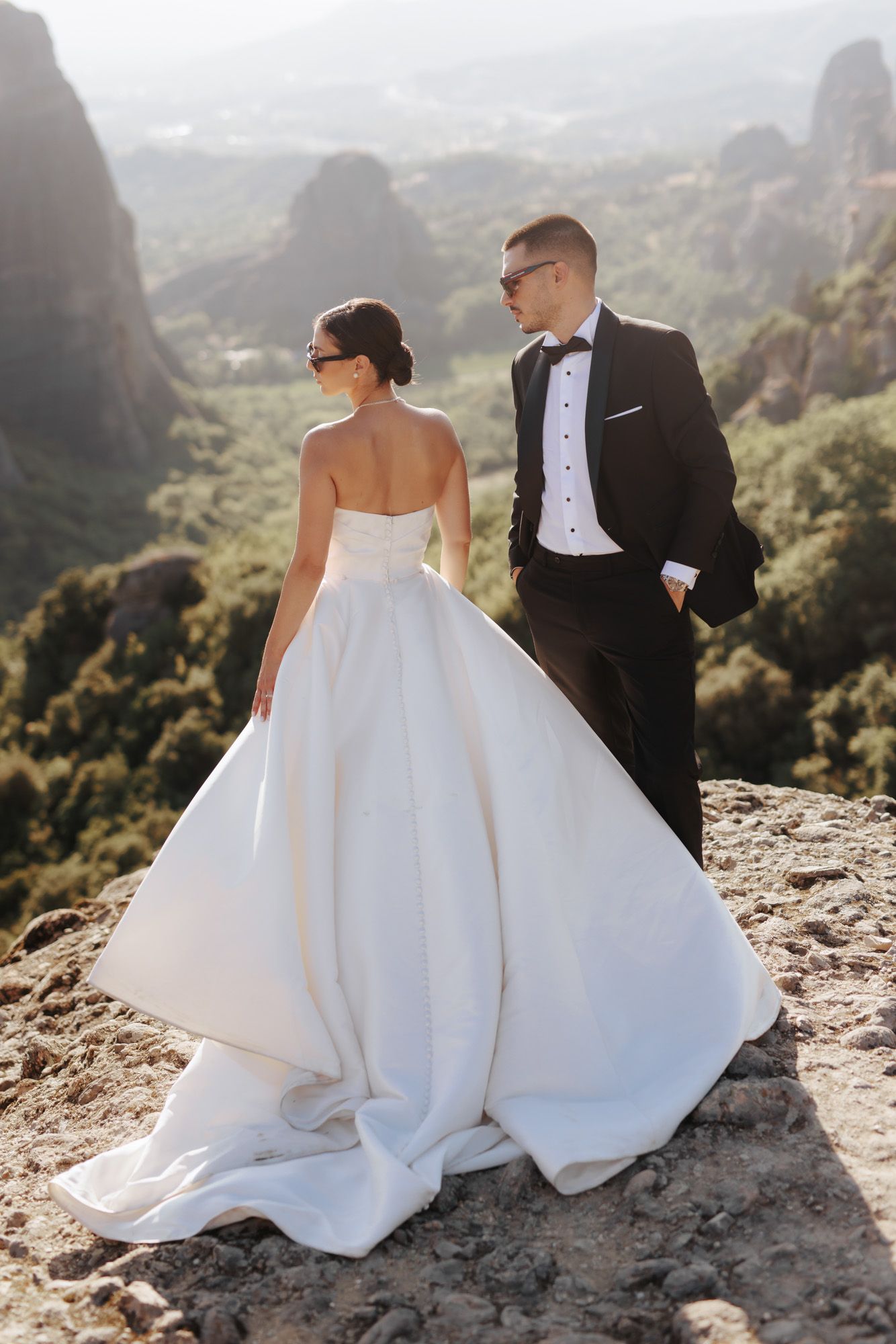 Bride and groom standing on rocky cliff with stunning mountain valley views, bride in strapless white gown, groom in black tuxedo.