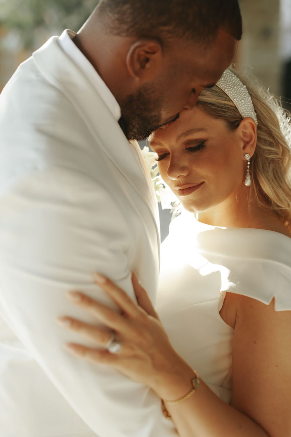 A bride and groom share an intimate moment, glowing in warm sunlight, as she wears a white gown and sparkling headpiece.