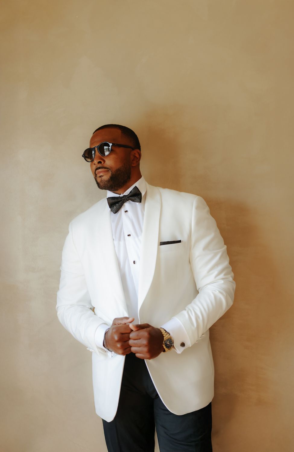 A stylish man in a white tuxedo jacket, black bow tie, and sunglasses poses confidently against a beige wall.