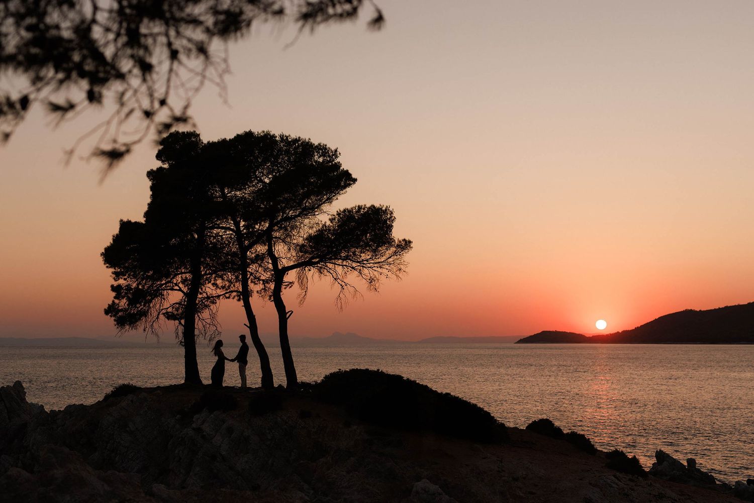 Silhouette of couple holding hands under pine trees at rocky coastline during vibrant orange sunset over calm sea.