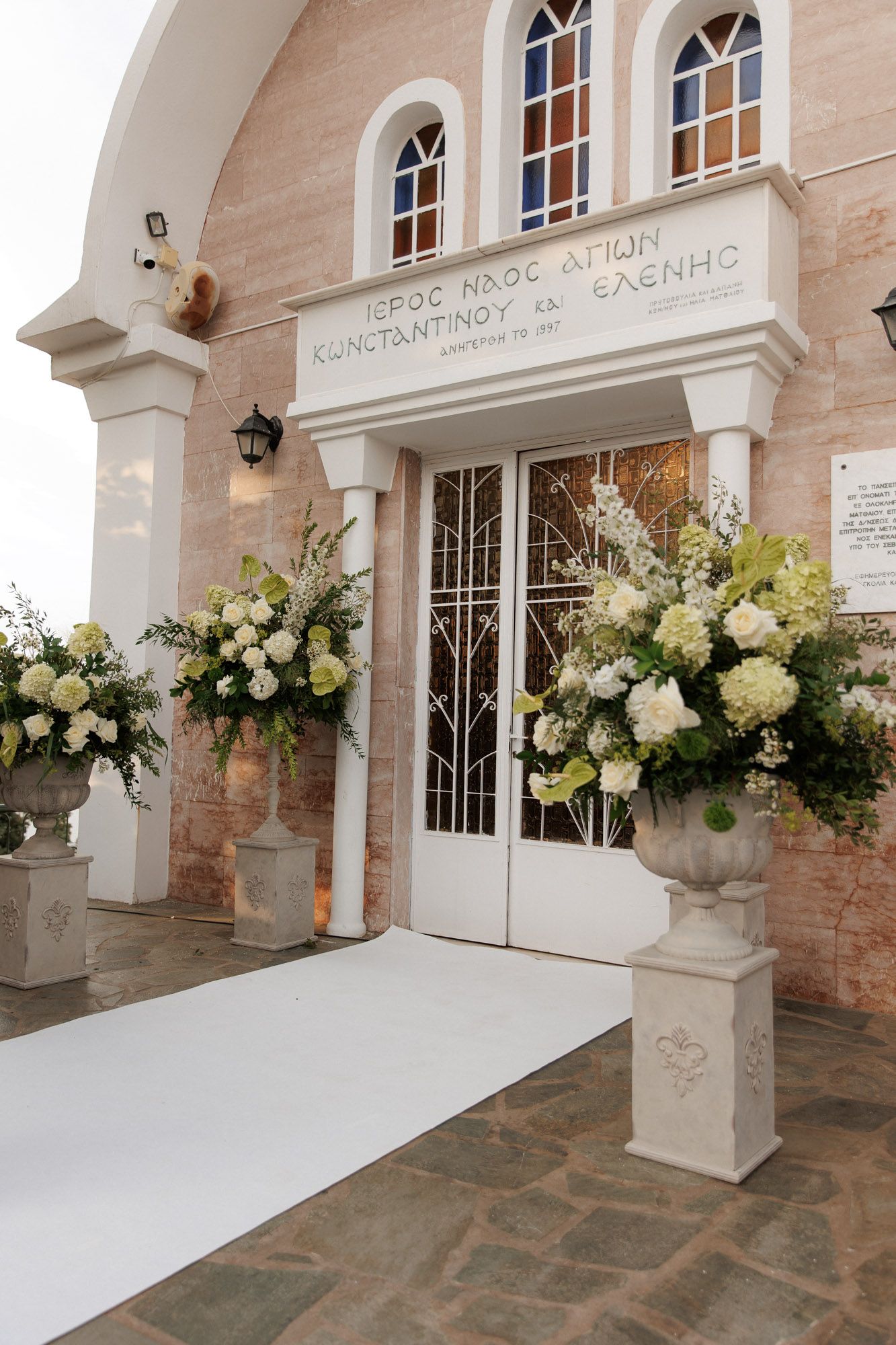 White floral arrangements in stone urns frame the entrance of a Greek Orthodox church with a white carpet aisle.