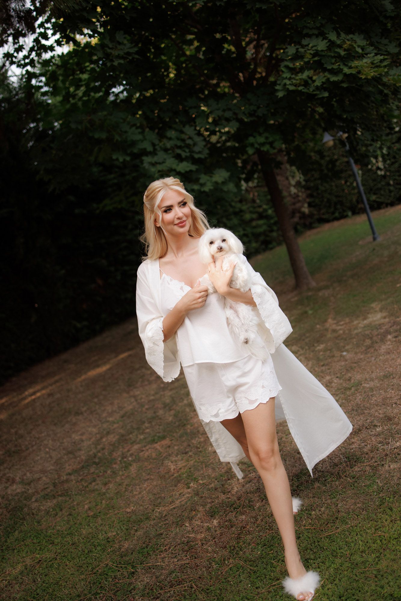 Smiling blonde woman in white silk robe and shorts holding a small white fluffy dog in a garden.