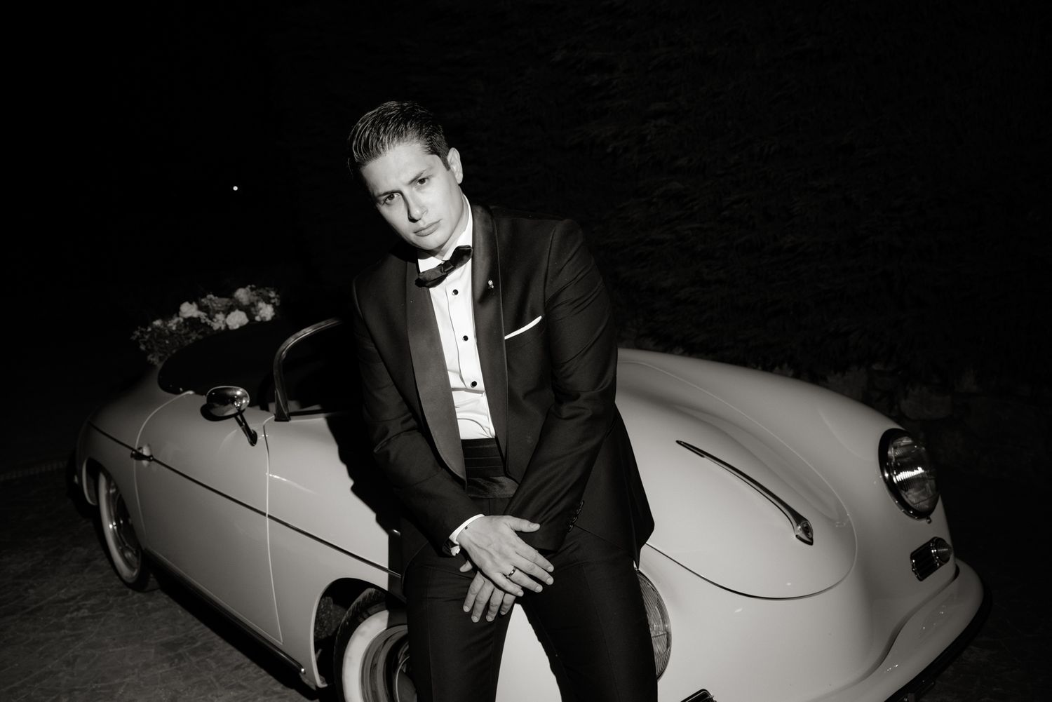 Groom in black tuxedo posing beside vintage cream convertible car in black and white.