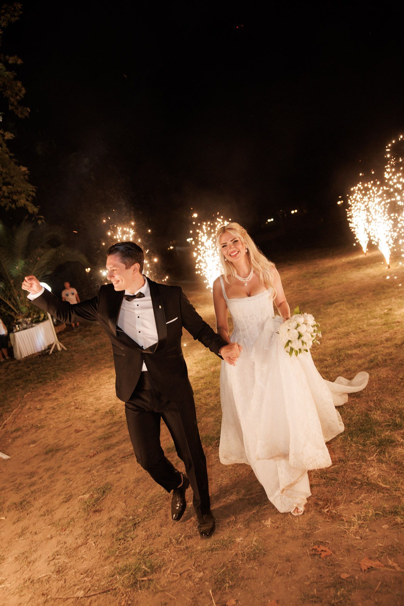 Bride and groom running through sparkling fireworks display at night wedding.