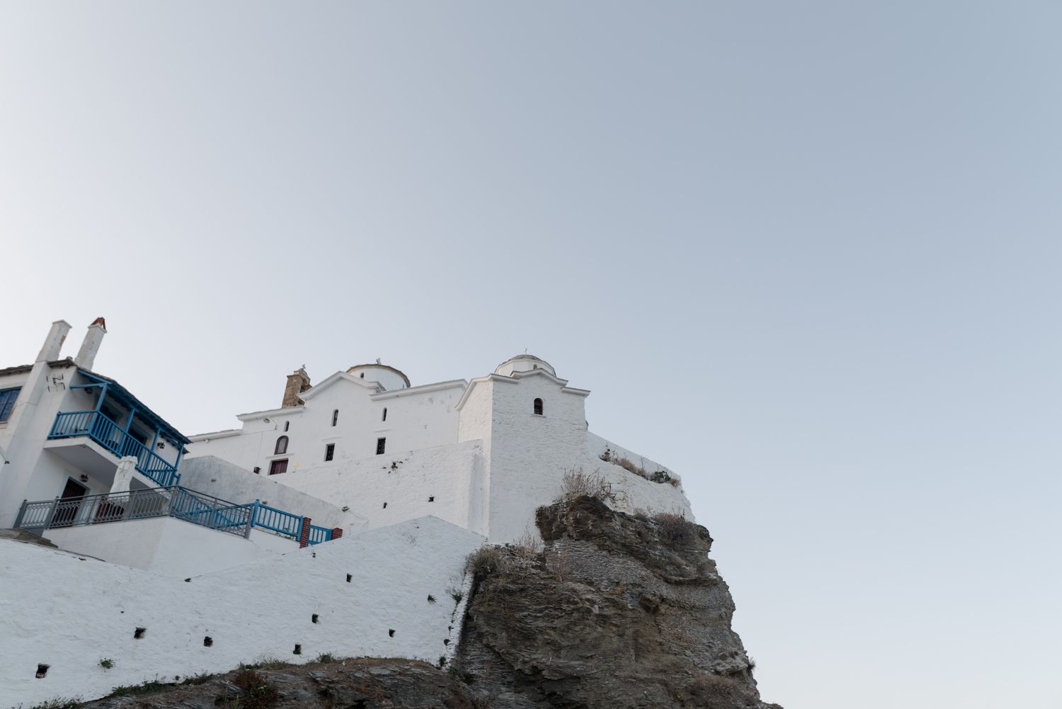 White-washed Greek island building perched dramatically on rocky cliff against clear sky.