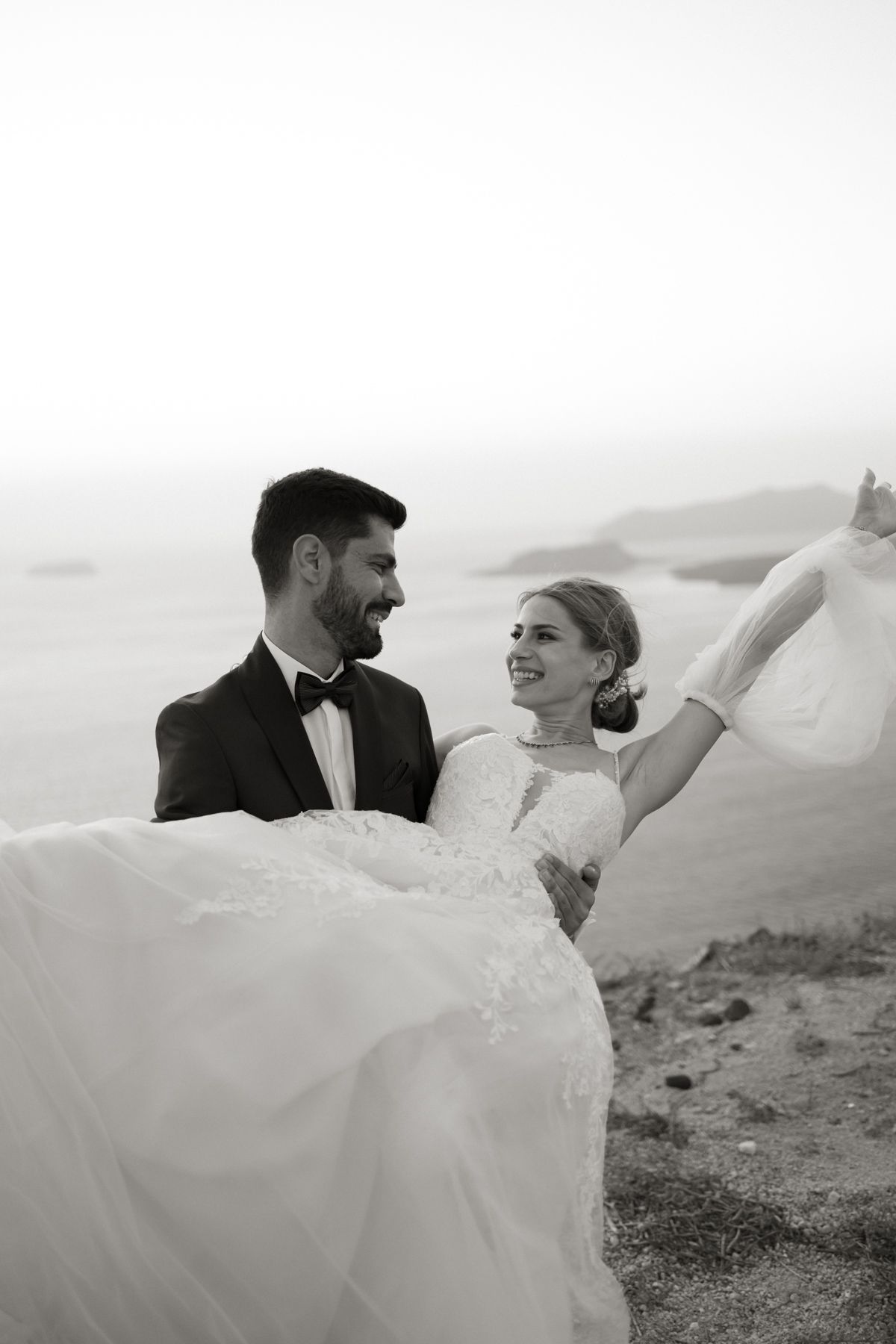 Black and white photo of smiling bride and groom embracing on beach, wedding dress flowing in the breeze.