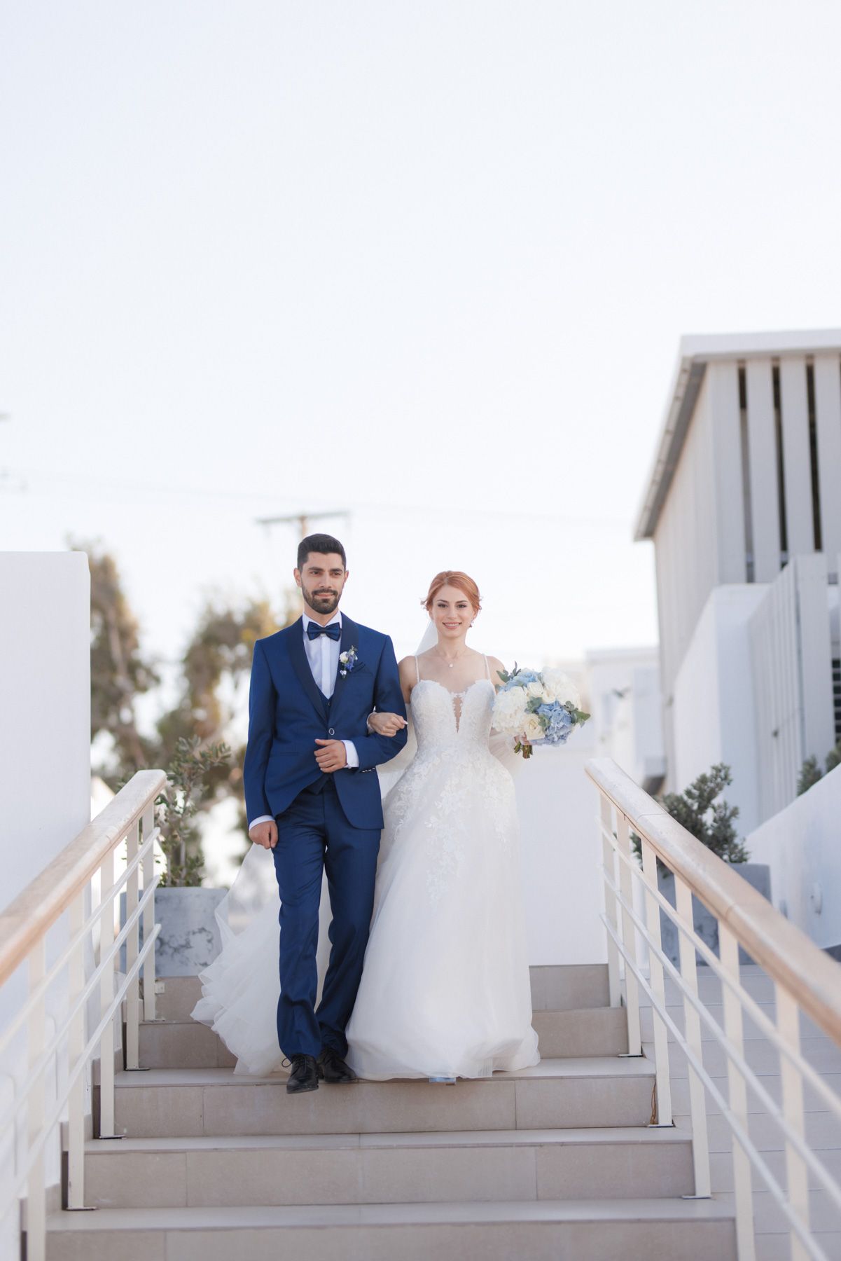 Bride and groom descending white steps at Santorini wedding venue, groom in navy tuxedo.