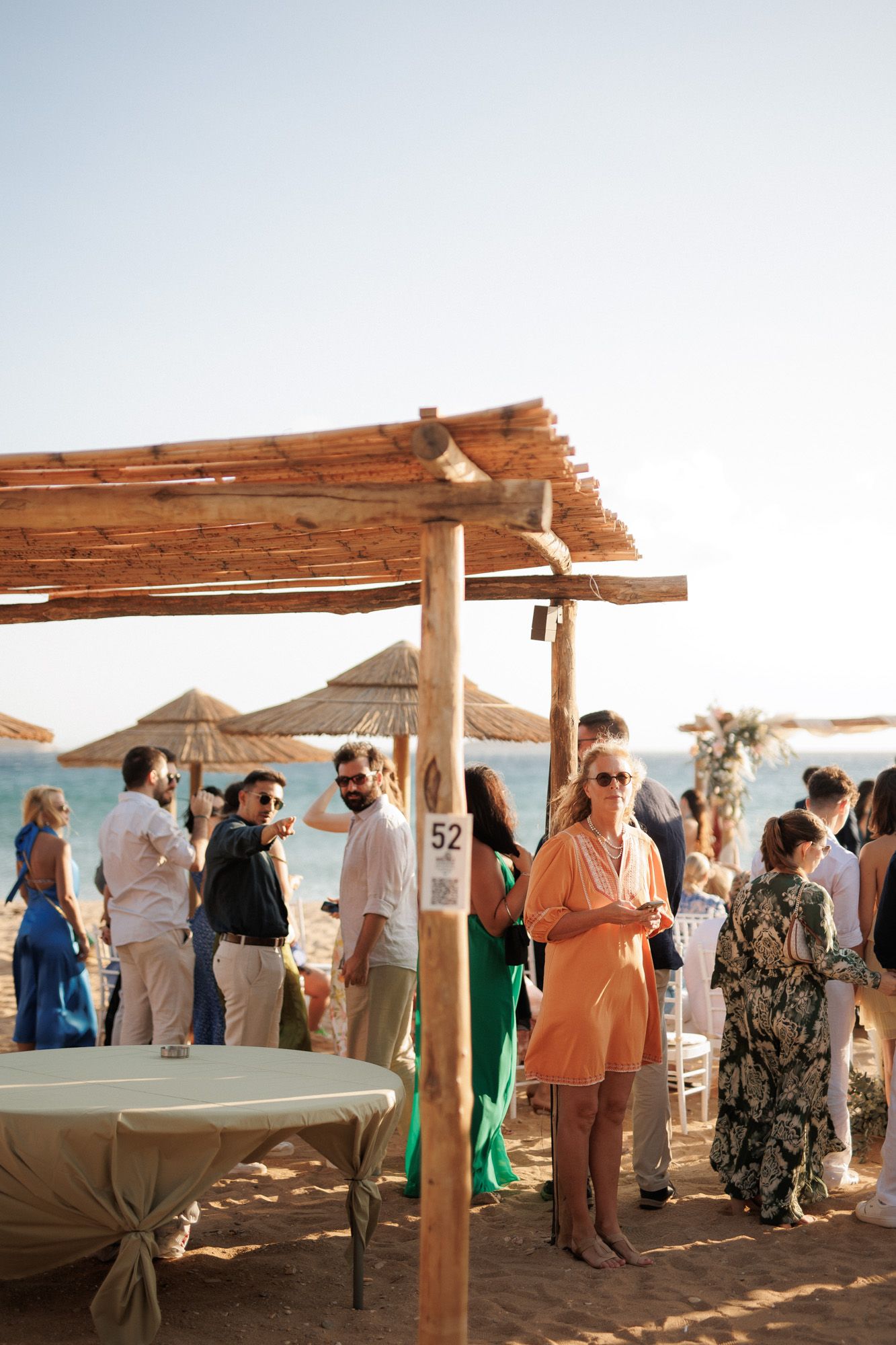 Guests mingle at a beach wedding reception under rustic wooden pergolas with straw umbrellas at sunset.