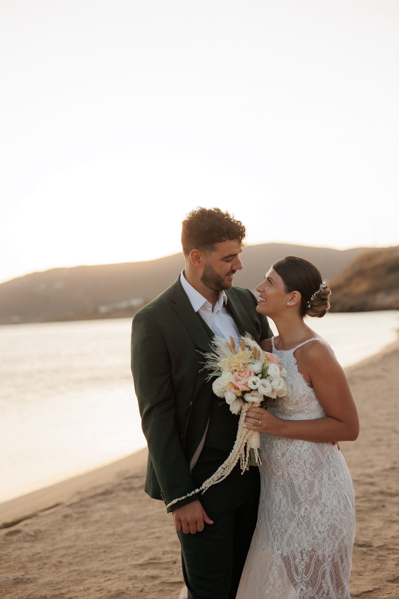 Bride and groom share romantic moment holding bouquet at golden hour on sandy beach.