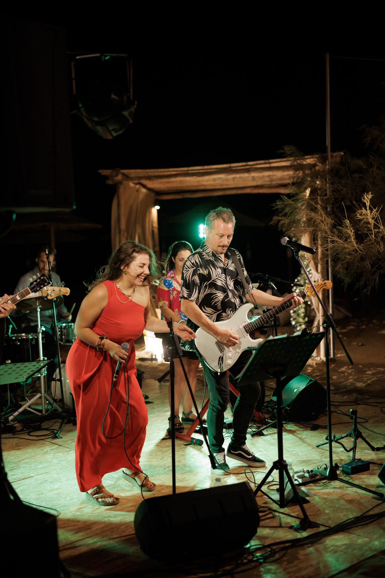 Live band performing outdoors at night with woman in red dress and guitarist on stage.