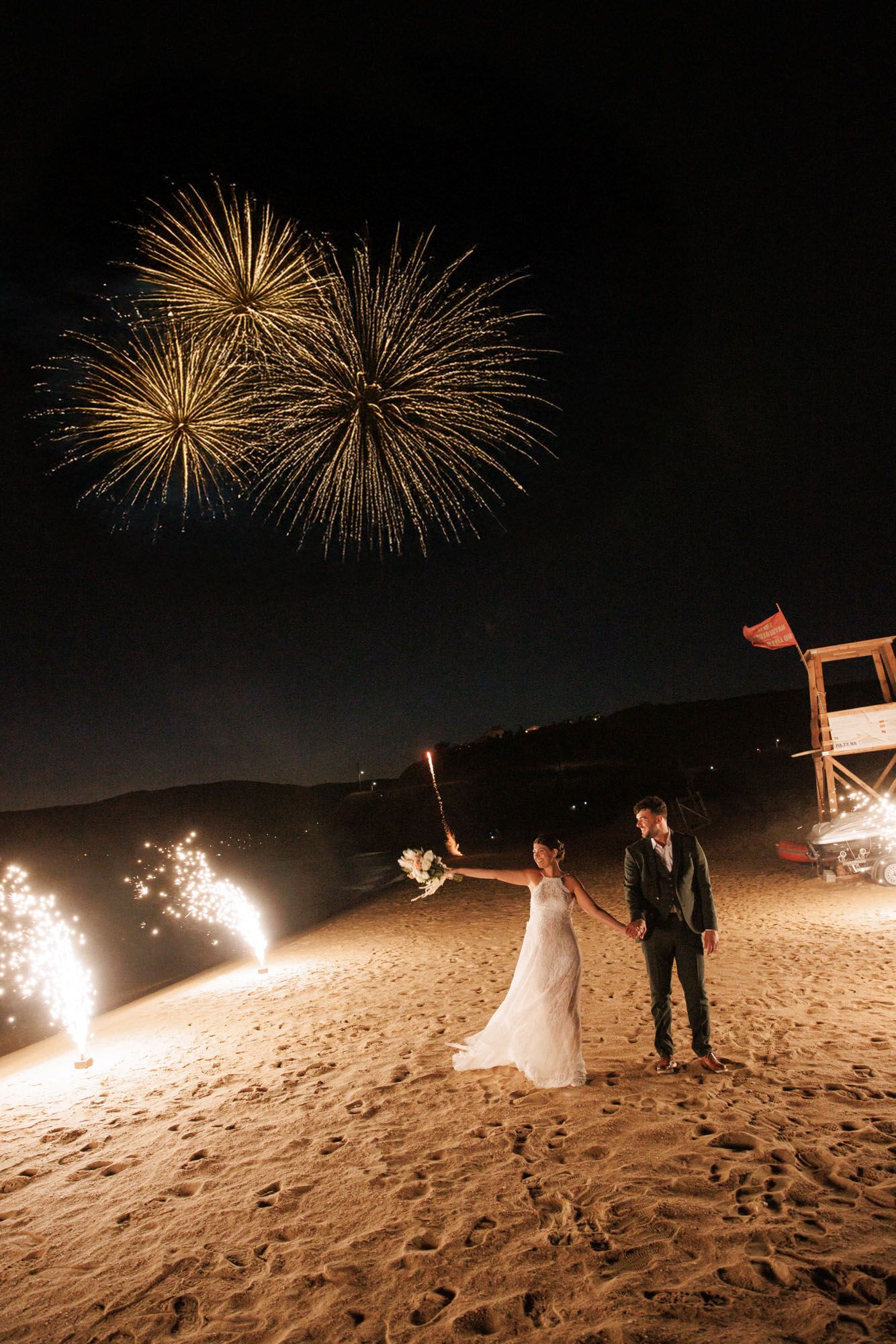 Bride and groom stand on beach at night with sparkling fireworks display overhead.