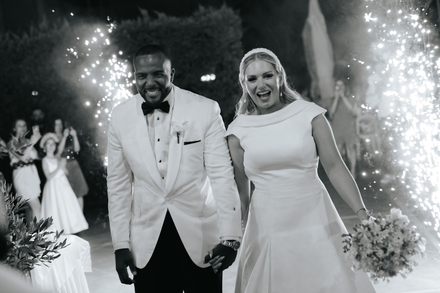 Black and white photo of a joyful couple exiting their wedding ceremony amid sparkling fireworks, bride holding bouquet.
