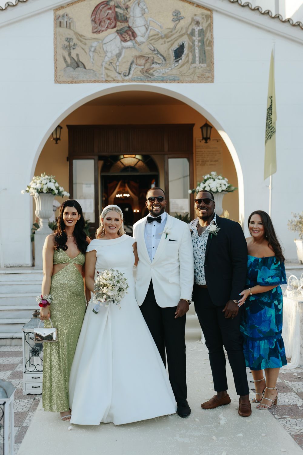 Wedding party posing outside a Greek Orthodox church, bride in white gown holding bouquet with guests in formal attire.