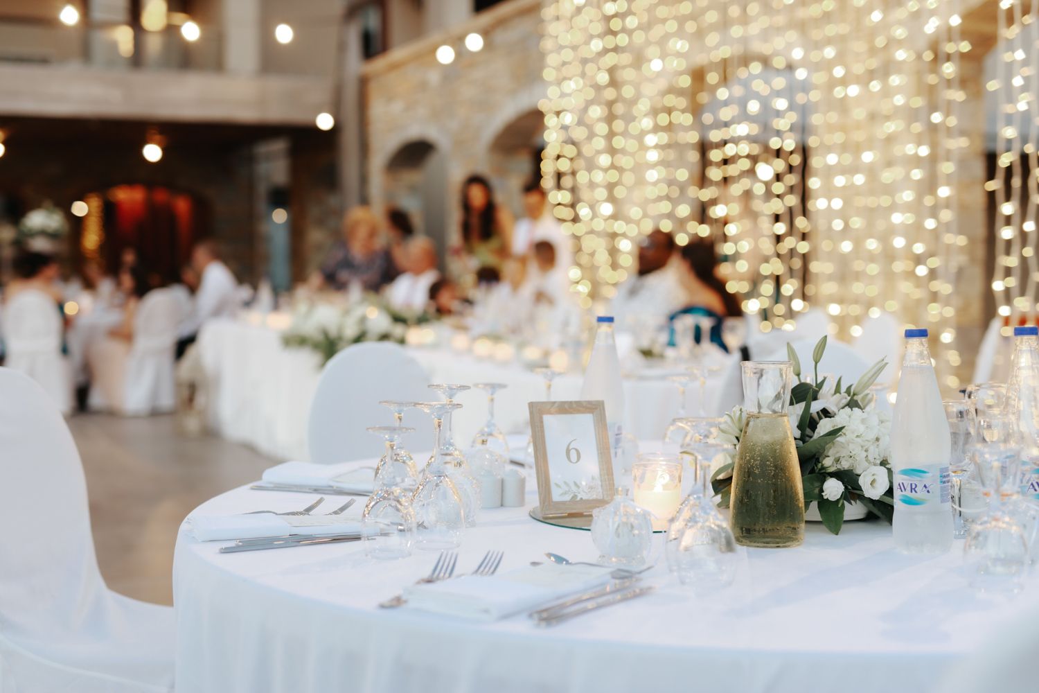 Elegant wedding reception with white tablecloths, glassware, floral centerpieces, and warm fairy lights in background.