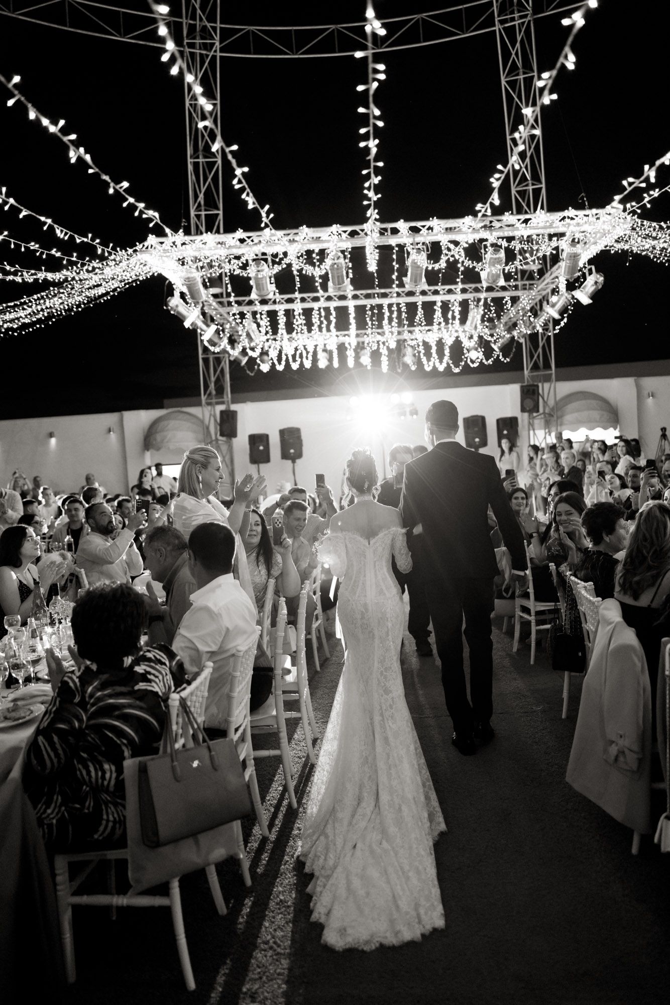 Black and white photo of bride and groom walking into wedding reception under sparkling chandelier lights at night.
