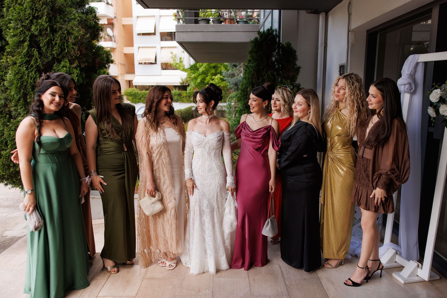 Bride in white lace gown poses with bridesmaids in colorful elegant dresses outdoors at a wedding celebration.