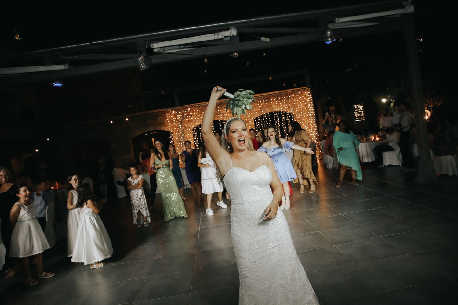 Bride in white gown tossing bouquet to guests on dance floor at elegant wedding reception.