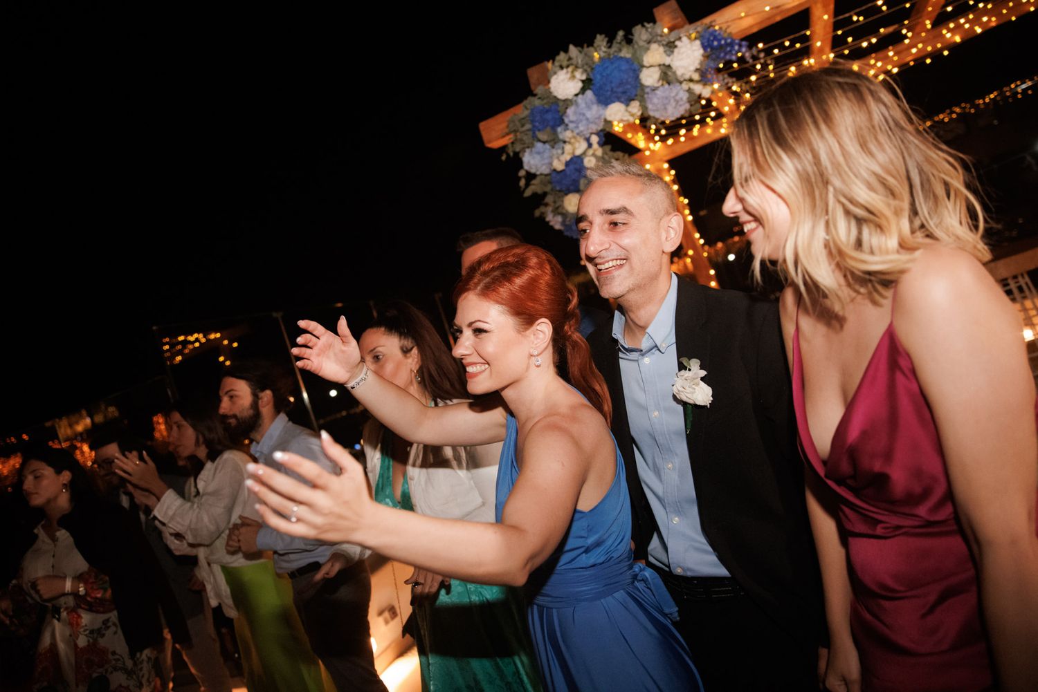 Wedding guests taking a selfie together at an outdoor evening reception with festive string lights in background.