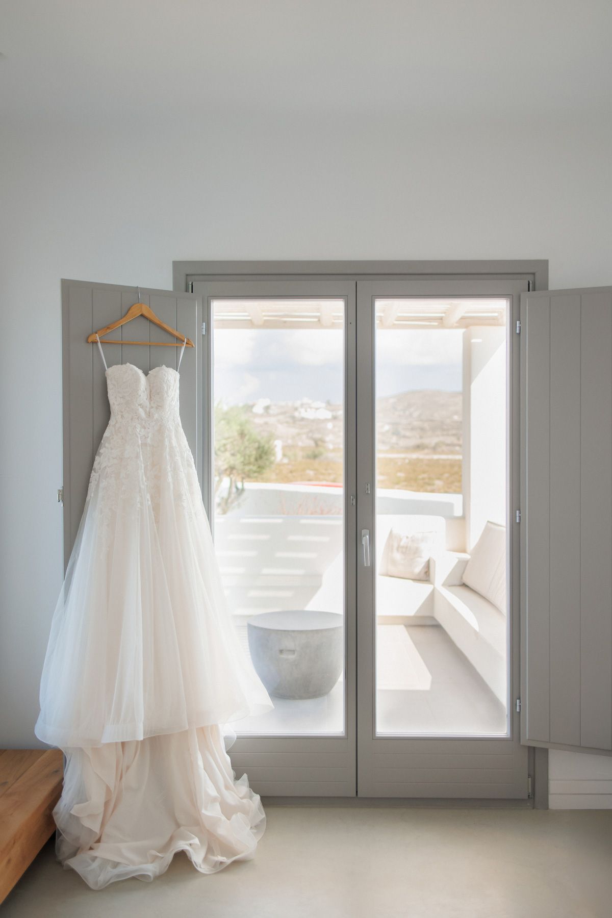 Elegant white lace wedding gown hanging on a wooden hanger in front of French doors with a scenic view.