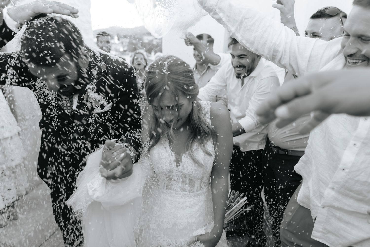 Black and white photo of bride and groom being showered with rice by wedding guests outdoors.